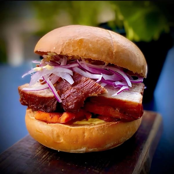 A Peruvian chicharrón sandwich with crispy pork belly, sweet potato, and red onion on a bun.