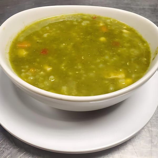 A white bowl of vibrant green Peruvian soup with vegetables and grains on a white plate.